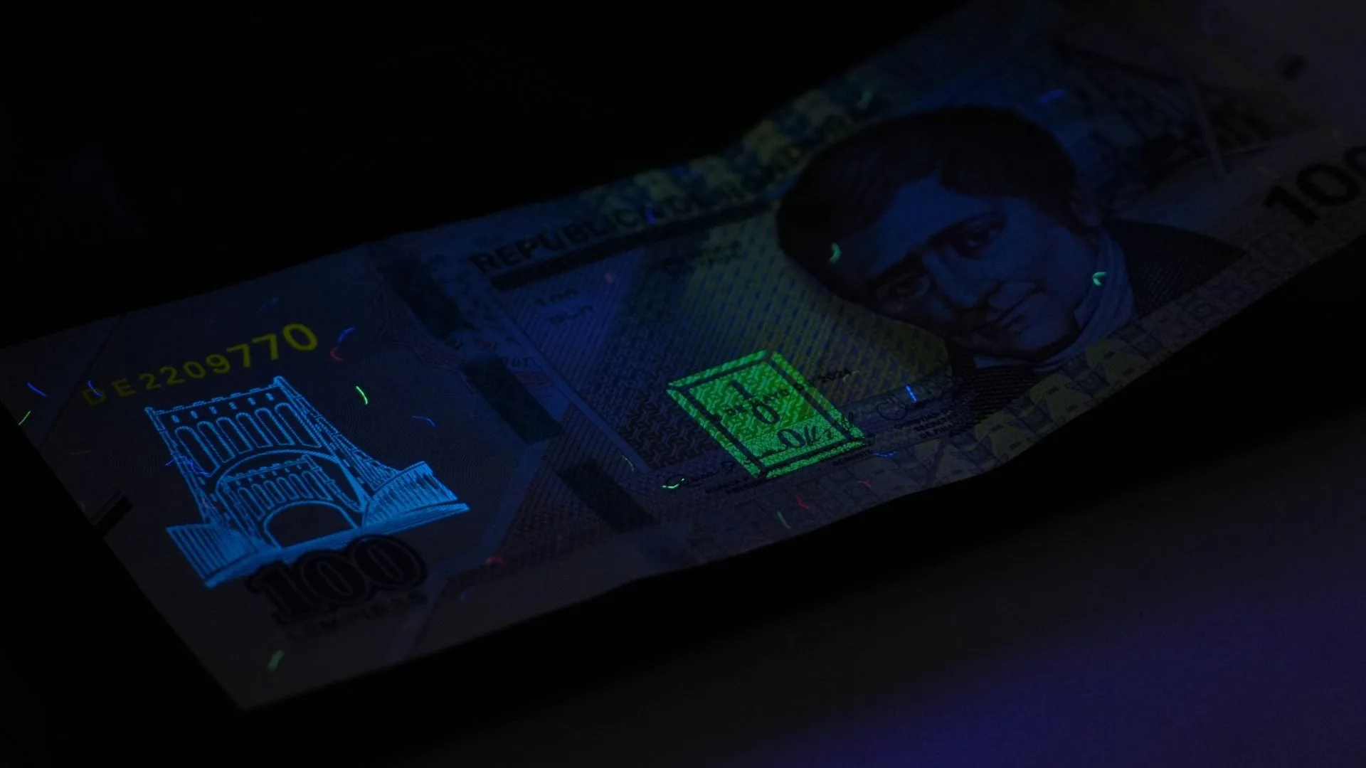 Billete de 100 lempiras bajo la luz ultravioleta, con sus fibras y marcas de seguridad fluorescentes.