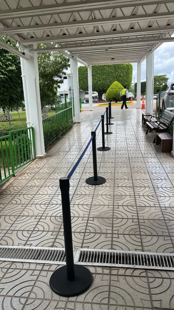 Postes separadores de fila con cinta azul a lo largo de un pasillo techado al aire libre.