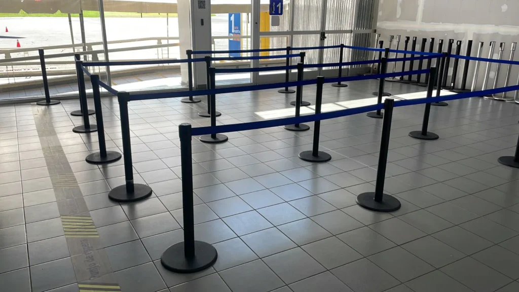 Postes separadores de fila con cinta azul formando una fila en zigzag junto a unos ventanales.