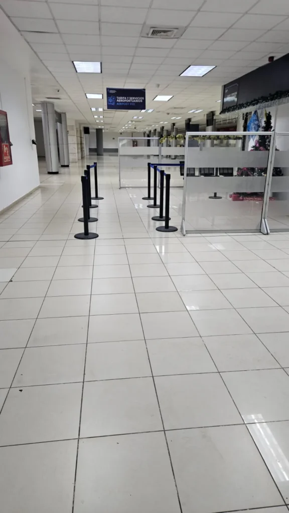 Postes separadores de fila con cinta azul junto a una mampara de vidrio en el vestíbulo de un aeropuerto.