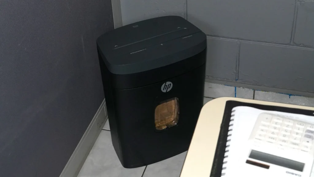 Trituradora de papel HP MC145 en la esquina de una oficina, junto a un escritorio con calculadora.